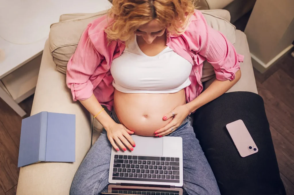 A pregnant woman sits on a couch, typing on a laptop with her one hand resting on her belly